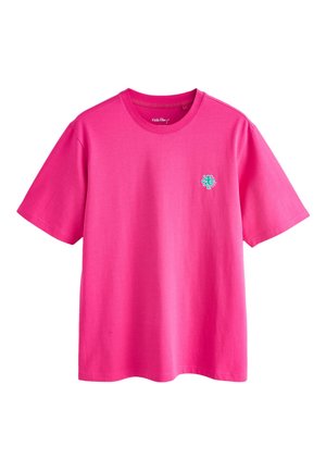 Junger Mann steht mit den Händen hinter dem Rücken, trägt ein knallpinkes T-Shirt, helle Jeansshorts und graue Sneakers vor einfarbig beigem Hintergrund.