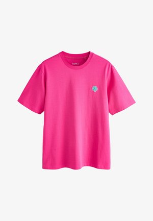 Junger Mann steht mit den Händen hinter dem Rücken, trägt ein knallpinkes T-Shirt, helle Jeansshorts und graue Sneakers vor einfarbig beigem Hintergrund.