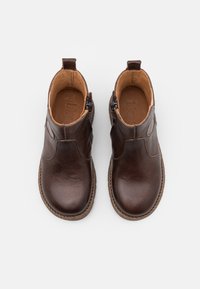 Pár tmavě hnědých kožených kotníkových bot s bočními zipy, zaoblenými špičkami a béžovým podšívkou na bílém pozadí.