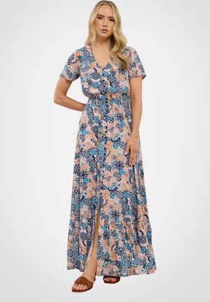 Kvinde iført en farverig blomster maxi-kjole med korte ærmer og knappet frontslids, kombineret med brune sandaler med åben tå, stående mod hvid baggrund.