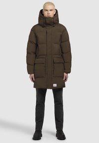 Brun polstret parka med høj krave og justerbar hætte, der har to forlommer og et logo-patch i kanten. Bæres over sorte bukser og støvler.