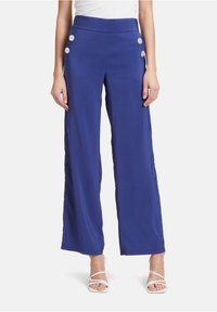 Breda byxor i ljusblått tyg med en slät textur, med hög midja och dekorativa knappar på sidorna.