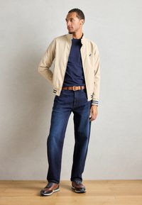 Kaki bomberjakke med marineblå striper, marineblå t-skjorte, blå denimjeans, brun belte og brune sko med grå detaljer, som står mot en grå vegg.