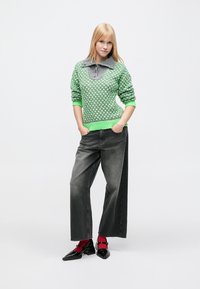 Pull vert tricoté avec col gris et motif texturé, associé à un jean gris à jambes larges et des chaussures noires avec des accents rouges.