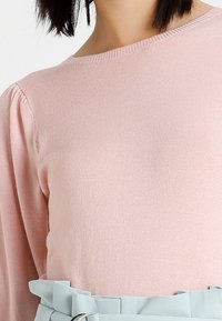 Pull en maille rose clair avec un col rond et des manches longues bouffantes, associé à une ceinture bleue douce avec un détail de boucle.