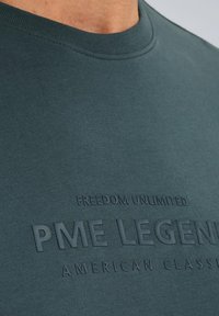 Mørkegrøn bomulds t-shirt med rund hals og hævet tekstgrafik: "FREEDOM UNLIMITED PME LEGEND AMERICAN CLASSIC." Blød struktur.
