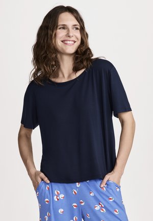Smiling woman with wavy hair wears a loose navy blue T-shirt and light blue shorts with red and white beach ball prints, hands in pockets.