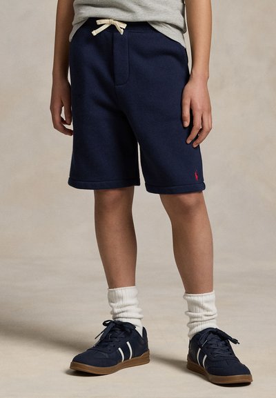Kind trägt marineblaue Shorts mit Kordelzug, weiße Socken und marineblaue Sneakers mit weißen Streifen, steht auf einem beigen Boden.