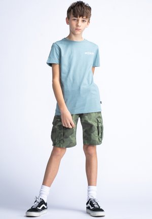 T-shirt en coton bleu clair avec "PETER" imprimé, accompagné de shorts cargo camouflage verts. Baskets noir et blanc avec des chaussettes blanches mi-mollet.