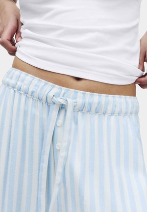 Personne levant une chemise blanche, portant un pantalon à rayures verticales bleu clair et blanches avec une taille élastique et un cordon de serrage.