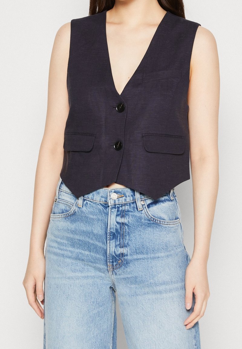 Veste noire sans manches à boutons avec deux poches avant, portée sur un jean en denim bleu clair taille haute.