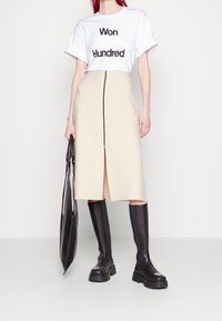 Camiseta blanca corta con texto negro, falda beige con abertura frontal y cremallera negra, combinada con botas negras brillantes hasta la rodilla y un bolso negro.