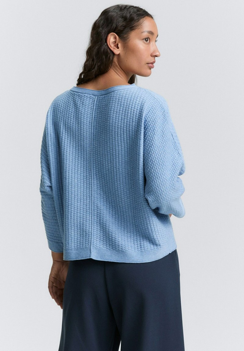Jasnoniebieski sweter o splocie waflowym, w luźnym kroju, skróconej długości, z opadającymi ramionami i szerokim dekoltem. Posiada teksturowaną powierzchnię.