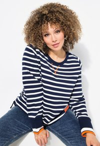 Chemise à rayures bleu marine et blanche à manches longues avec un accent orange au niveau du col et des poignets, associée à un jean en denim, tissu texturé.