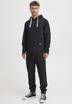 Schwarzer Zip-Overall mit Kapuze, vorn mit Taschen, beigen Kordeln und einem kleinen Lederpatch. Rippbündchen an den Ärmeln und Beinen.