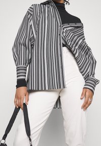 Camisa de rayas en blanco y negro de gran tamaño con mangas globo y cierre de botones, combinada con pantalones blancos ajustados y un accesorio de tirante negro.