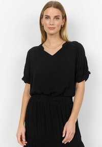 Čierna blúzka s výstrihom do V, krátke volánové rukávy, vyrobená z hladkého materiálu, v kombinácii s plisovanou čiernou sukňou. Jednoduchý a elegantný dizajn.