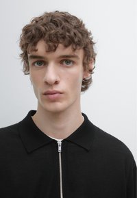 Černé polo tričko na zip s límečkem, vyrobené z hladké látky. Model má kudrnaté vlasy a stojí před jednobarevným pozadím.