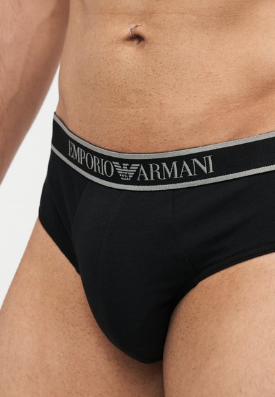 Ανδρικός κορμός που φοράει μαύρο εσώρουχο Emporio Armani με ορατό λογότυπο στη ζώνη και ασημένιο δαχτυλίδι στον ομφαλό.
