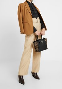 Žena v camel saku přes černou košili a béžovými kalhotami s vysokým pasem, držící černou kabelku s hadí texturou, v hnědých špičatých botách.