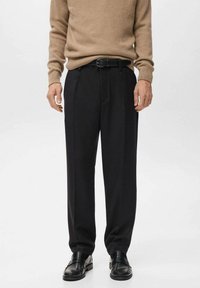 Pantalones negros con un ajuste entallado, con pliegues frontales y una textura suave, combinados con un suéter de punto beige y zapatos negros.