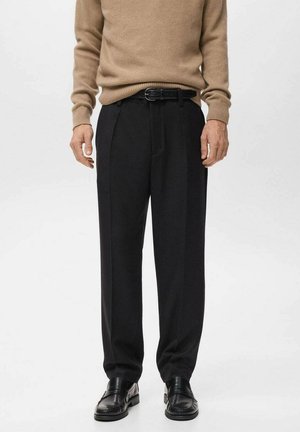 Pantalon noir à coupe ajustée, avec des plis à l'avant et une texture lisse, associé à un pull en tricot beige et des chaussures noires.