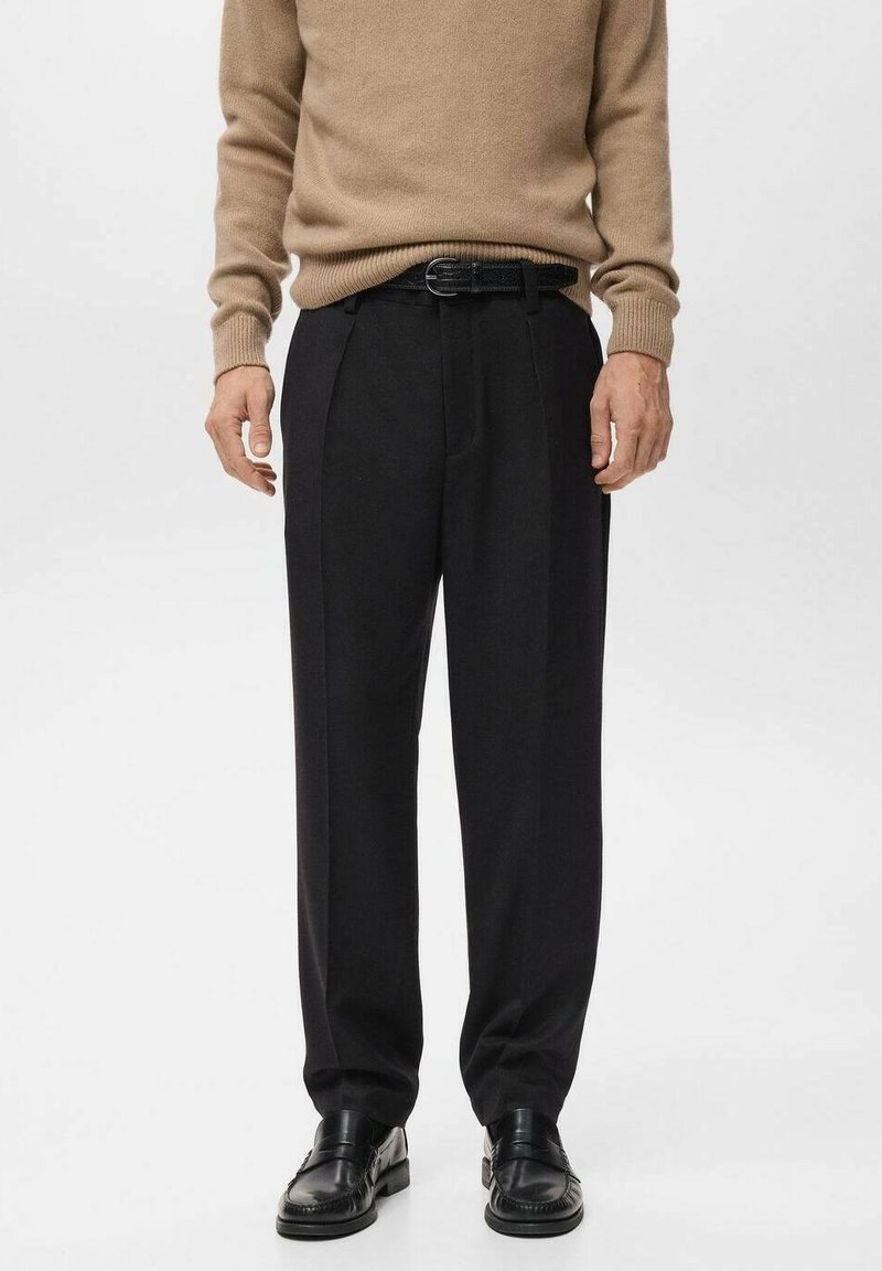 Pantalones negros con un ajuste entallado, con pliegues frontales y una textura suave, combinados con un suéter de punto beige y zapatos negros.