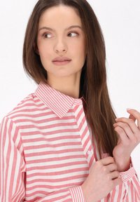 Różowo-biała koszula w paski z klasycznym kołnierzykiem, zapięciem na guziki i długimi rękawami z akcentowanymi paskami przy mankietach. Gładka tekstura materiału.