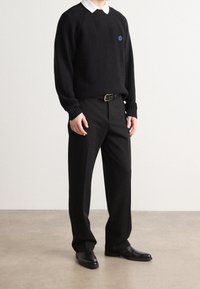 Pull noir côtelé avec une chemise blanche à col, assorti à un pantalon noir et une ceinture. Des chaussures formelles noires complètent la tenue.