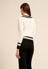 Donna di spalle che indossa un maglione crema con polsini e orlo a righe nere e dorate, abbinato a pantaloni neri, su uno sfondo semplice.