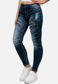 Jeans skinny blu in denim con molte tasche decorative con cerniera, lavaggio sbiadito, aree strappate e vestibilità affusolata, abbinati a sneakers bianche.