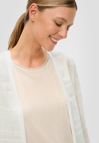 Leichter, weißer Cardigan mit subtiler gestreifter Textur über einem weichen, schimmernden beigefarbenen Oberteil. Beide Kleidungsstücke verfügen über entspannte Designs und runde Ausschnitte.