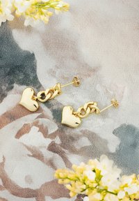 Guldörhängen i hjärtform med en texturerad kedja som förbinder hjärtana, avbildade mot en blommönstrad bakgrund.