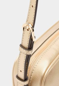 Gouden metallic leren handtasriem met een zwarte rand, voorzien van een gouden sluiting en zichtbare stiksels.