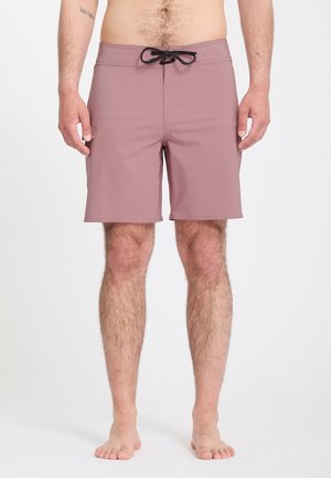 Homme torse nu portant un short de bain mauve avec un cordon noir, debout pieds nus sur un fond blanc uni.