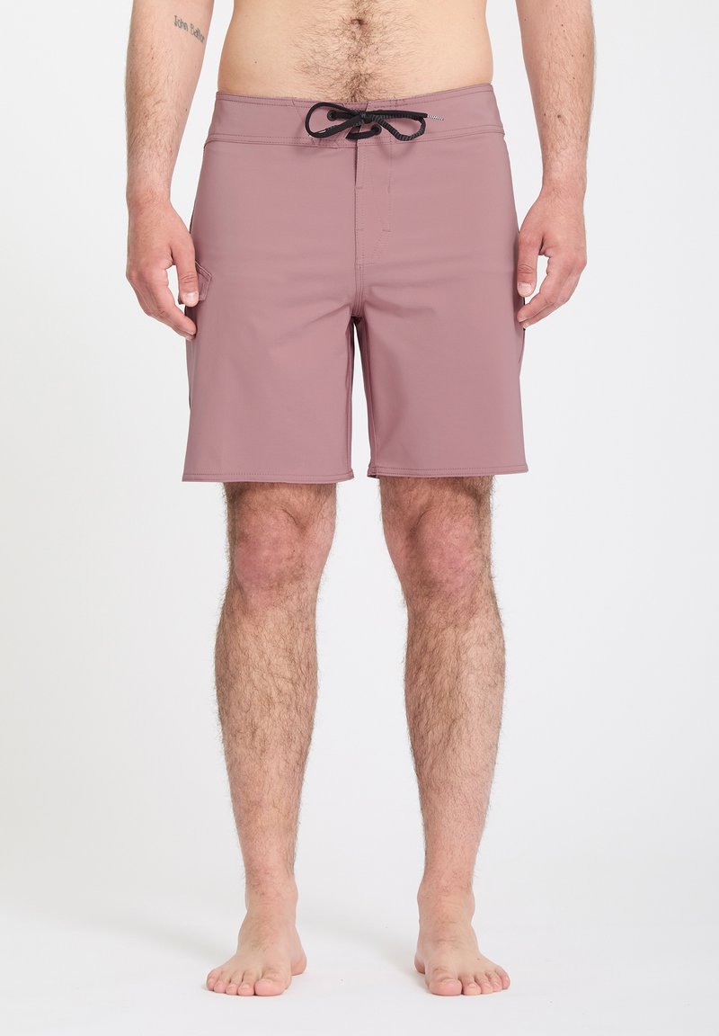 Homme torse nu portant un short de bain mauve avec un cordon noir, debout pieds nus sur un fond blanc uni.