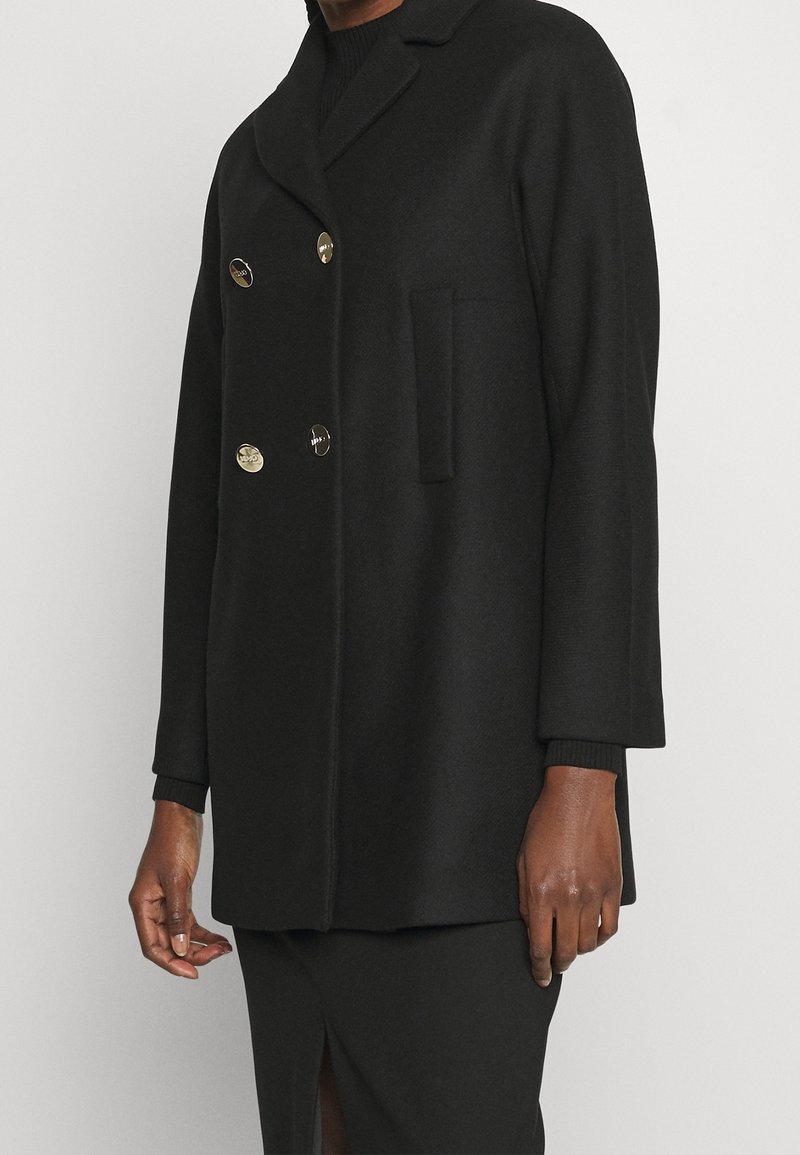 Manteau noir croisé à double boutonnage avec quatre boutons dorés porté sur un haut à manches longues noir et une jupe noire fendue sur le devant.