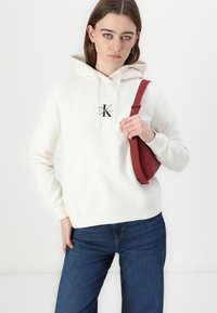 Balta gobtuvė su priekinėmis logotipais, su raišteliu reguliuojamu gobtuvu, išgaubtais rankogaliais, derinama su tamsiai mėlynomis džinsomis ir raudona per petį nešama rankine.