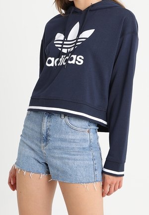 Personne portant un sweat à capuche Adidas bleu marine avec logo et bordure blanche, associé à un short en jean bleu clair taille haute aux bords effilochés.