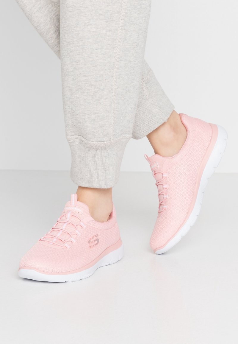 Personne portant un pantalon gris clair et des baskets Skechers roses avec des semelles blanches, debout sur un sol et un fond blancs.