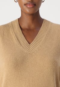 Hellbrauner, gestrickter V-Ausschnitt-Pullover mit ribbeligem Kragen, der eine weiche Textur und subtile tonale Unterschiede aufweist. Keine sichtbaren Muster oder Akzente.
