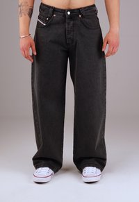 Wide-leg black denim jeans with a faded finish, featuring five pockets and metal hardware accents. Paired with white sneakers.