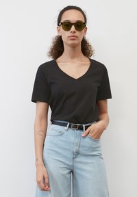 Svart V-hals t-skjorte laget av bomull, parret med lyseblå høytlivsjeans og et svart belte. Modellen har på seg grønnfargede solbriller.