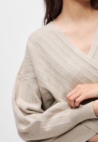 Femme portant un pull cache-cœur beige en tricot côtelé à manches longues, tenant délicatement une manche avec les mains croisées.
