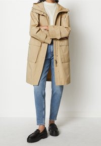 En person klädd i en beige huppjacka, med armarna i kors, kombinerad med ljusblå jeans och svarta kraftiga loafers, stående mot en helt vit vägg.