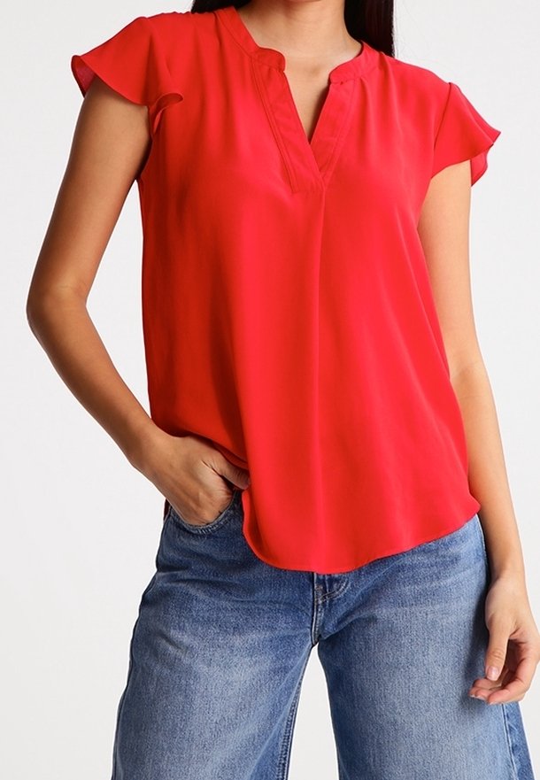 Blouse rouge à manches courtes avec un décolleté en V, faite de tissu léger ; texture lisse et coupe ample. Associée à un jean bleu.