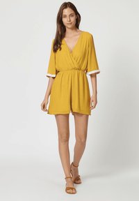 Mono amarillo con diseño envolvente, mangas cortas y cintura elástica. Presenta adornos de encaje blanco en los bordes de las mangas y pantalones cortos con plisados.