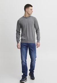 Grå, langermet genser i jevn strikk, kombinert med blå denimjeans. Modellen har på seg svarte joggesko og står mot en ensfarget bakgrunn.