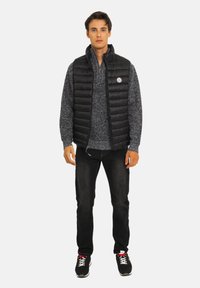 Doudoune noire sans manches avec coutures horizontales, pull en maille gris en dessous, jean noir et baskets noires avec des accents rouges. Le mannequin est debout, droit.