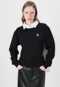 Svart sweatshirt med rund halsringning, med en liten grafisk tryck på bröstet. Kombination med en vit skjorta med krage under.